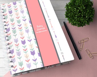 May include: A pink and white baby journal with a gray and white patterned cover. The cover has a pink stripe with the text "Baby Journal" and "For Riley & Emma". The journal has a spiral binding and is decorated with arrows.