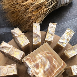 May include: A close-up of a wooden cutting board with multiple bars of handmade soap. The soap is a light brown color with swirls of white and gold flakes. The soap is arranged in a circular pattern on the board.