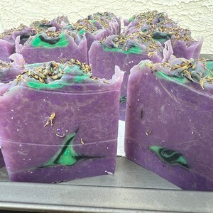 May include: A close-up of several bars of handmade soap. The soap is purple with green and black swirls and has dried lavender flowers on top.