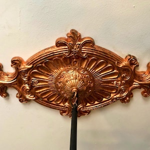 May include: A gold-colored metal ceiling medallion with an ornate design. The medallion has a central circular design with a smaller circular opening in the center.