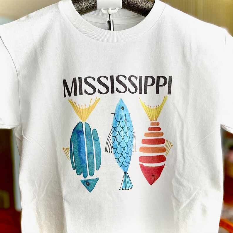 K&ouml;nnte beinhalten: Wei&szlig;es T-Shirt mit dem Wort "MISSISSIPPI" in Schwarz bedruckt. Unter dem Wort sind drei stilisierte Fische gedruckt, ein blauer, ein gr&uuml;ner und ein orange-roter.