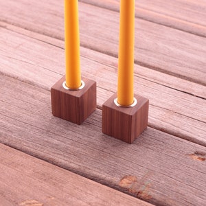 May include: Two wooden candle holders with yellow candles. The candle holders are square and made of dark wood. The candles are a bright yellow color. The holders are on a weathered wooden surface.