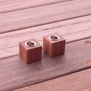 May include: Two square, wooden candle holders with a silver-colored metal insert. The candle holders are a warm brown color and sit on a weathered wooden surface.