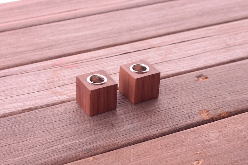 May include: Two square, wooden candle holders with a silver-colored metal insert. The candle holders are a warm brown color and sit on a weathered wooden surface.