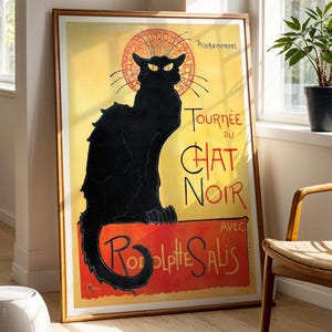 May include: A framed poster featuring a black cat silhouette with yellow eyes, set against a yellow and red background. The poster reads "Tournée du Chat Noir" with additional text. The artwork is displayed in a room with natural light and a wooden chair.