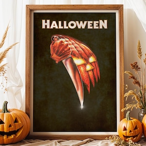 Peut inclure: Une affiche encadr&eacute;e avec le mot HALLOWEEN en lettres capitales blanches en haut. Le graphisme central repr&eacute;sente une main tenant un grand couteau argent&eacute;, dont la lame se transforme en une citrouille orange lumineuse sur fond sombre.