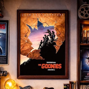May include: A framed poster for "The Goonies" movie, with a group of figures silhouetted against a sunset, hangs above a wooden desk. A director's chair, a shark figurine, a fedora, and a globe lamp are on the desk.