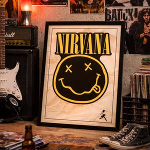 May include: A framed Nirvana poster with the band's name in yellow and a black smiley face logo. The image is set in a room with music memorabilia, a guitar, and a pair of high-top sneakers.