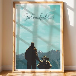Puede incluir: Un cuadro enmarcado con un paisaje de montaña y dos siluetas. El cuadro tiene un marco de madera clara y la palabra "Intouchables" en escritura cursiva. El fondo muestra un cielo azul, nubes blancas y montañas nevadas.