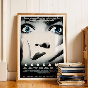 May include: A framed "Scream" movie poster, showing a close-up of a face with blue eyes and the film's title. The poster is in a wooden frame, leaning against a white wall. A stack of books is on the floor beside it.