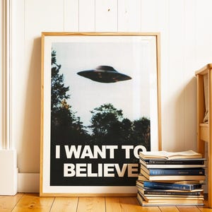 Könnte beinhalten: Gerahmter Kunstdruck mit einem Retro-Bild einer fliegenden Untertasse über Bäumen. Der Druck trägt die Worte "I WANT TO BELIEVE" in weißer Schrift auf schwarzem Hintergrund. Der Druck befindet sich in einem Holzrahmen und wird in Innenräumen ausgestellt.