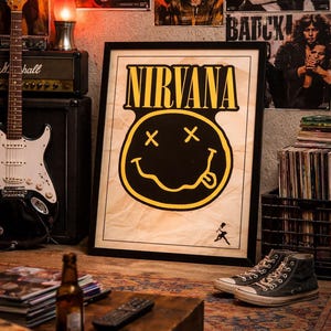 May include: Framed Nirvana poster featuring the band's logo, a yellow smiley face with crossed-out eyes and a tongue sticking out, set against a vintage paper background. The poster is in a room with a guitar, amplifier, and other rock memorabilia.