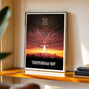 May include: Framed poster featuring a scene from the movie "Independence Day." The image shows a cityscape under an alien spacecraft, with the film's title at the bottom. The poster is in a gold frame and is displayed on a wooden shelf.
