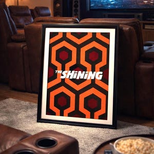 Può includere: Stampa artistica incorniciata con il motivo geometrico del film "The Shining". Il design è composto da esagoni arancioni, neri e bordeaux. Il titolo "THE SHINING" è visualizzato in bianco.