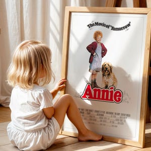 Könnte beinhalten: Gerahmtes Filmplakat für "Annie" mit dem Titel in Rot, einem Mädchen in roter Jacke und Kleid und einem Hund. Das Poster befindet sich in einem hellen Holzrahmen, und ein kleines Kind sitzt daneben auf dem Boden.
