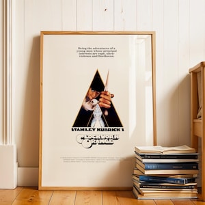 Op de afbeelding: Ingelijste filmposter voor Stanley Kubricks "A Clockwork Orange". De poster toont een centraal driehoekig beeld met een personage dat een wapen vasthoudt. De titel van de film staat eronder, met tekst erboven die de thema's van de film beschrijft. De lijst is van hout.