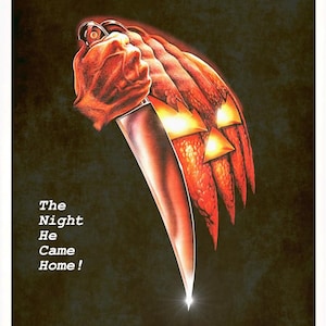 Peut inclure: Une affiche de film pour Halloween repr&eacute;sentant une grande citrouille orange sculpt&eacute;e avec des yeux et une bouche lumineux, tenue par une main agrippant un couteau argent&eacute;. Le texte indique HALLOWEEN, The Night He Came Home!, et les cr&eacute;dits.