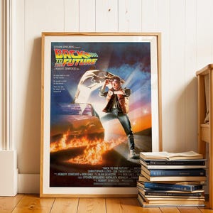 Może przedstawiać: Oprawiony plakat filmowy do filmu "Powrót do przyszłości" z tytułem filmu i sceną z płonącym samochodem. Plakat jest w drewnianej ramie i jest oparty o białą ścianę. Stos książek leży na podłodze.
