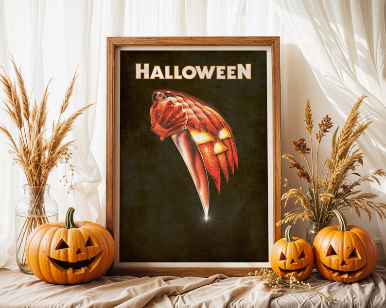 Peut inclure: Une affiche encadr&eacute;e avec le mot HALLOWEEN en lettres capitales blanches en haut. Le graphisme central repr&eacute;sente une main tenant un grand couteau argent&eacute;, dont la lame se transforme en une citrouille orange lumineuse sur fond sombre.