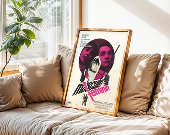 May include: A framed movie poster for "Masculin Féminin" by Jean-Luc Godard. The poster features a pink and black design with two faces in a circle and the title in pink. The poster is in a light brown wooden frame.