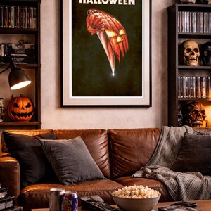 Peut inclure: Une affiche murale encadr&eacute;e pr&eacute;sentant le mot HALLOWEEN en lettres blanches au-dessus d'une illustration d'une main tenant un grand couteau de cuisine, dont la lame se transforme en citrouille illumin&eacute;e sur un fond sombre.
