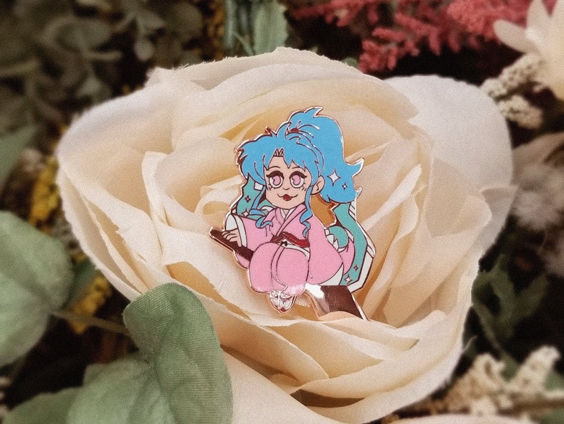 Pu&ograve; includere: Uno spillo in smalto blu e rosa con un personaggio dei cartoni animati con i capelli blu e un vestito rosa che cavalca una scopa. Lo spillo &egrave; posizionato su uno sfondo di rosa bianca.