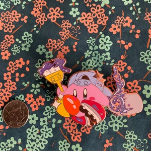 K&ouml;nnte beinhalten: Eine rosa und lilafarbene Emaille-Anstecknadel mit einer Comicfigur, die einen Pinsel und ein sternf&ouml;rmiges Objekt h&auml;lt. Die Figur tr&auml;gt einen blauen Hut mit wei&szlig;en Sternen. Die Anstecknadel hat eine glitzernde Oberfl&auml;che.