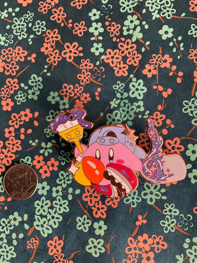 K&ouml;nnte beinhalten: Eine rosa und lilafarbene Emaille-Anstecknadel mit einer Comicfigur, die einen Pinsel und ein sternf&ouml;rmiges Objekt h&auml;lt. Die Figur tr&auml;gt einen blauen Hut mit wei&szlig;en Sternen. Die Anstecknadel hat eine glitzernde Oberfl&auml;che.