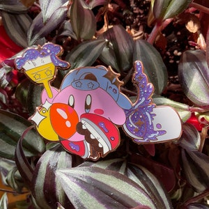 K&ouml;nnte beinhalten: Eine rosa und gelbe Emaille-Anstecknadel mit einer Comicfigur mit einem blauen Hut und einem Pinsel. Die Figur h&auml;lt einen gelben Stern und einen Pinsel mit lila und blauen Farbflecken. Die Anstecknadel hat ein glitzerndes Finish.