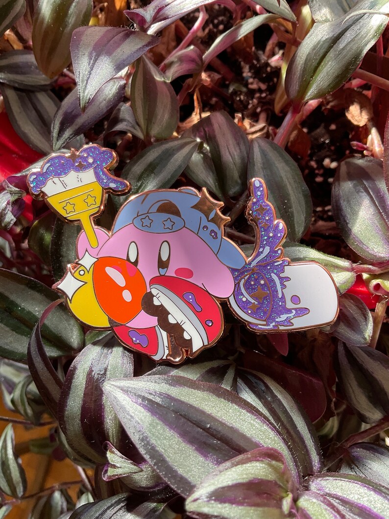 K&ouml;nnte beinhalten: Eine rosa und gelbe Emaille-Anstecknadel mit einer Comicfigur mit einem blauen Hut und einem Pinsel. Die Figur h&auml;lt einen gelben Stern und einen Pinsel mit lila und blauen Farbflecken. Die Anstecknadel hat ein glitzerndes Finish.