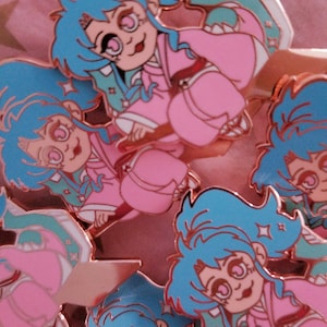 Pu&ograve; includere: Un set di cinque spille smaltate con un personaggio dei cartoni animati con capelli blu, vestiti rosa e occhi grandi. Le spille sono in varie pose e hanno una finitura metallica lucida.