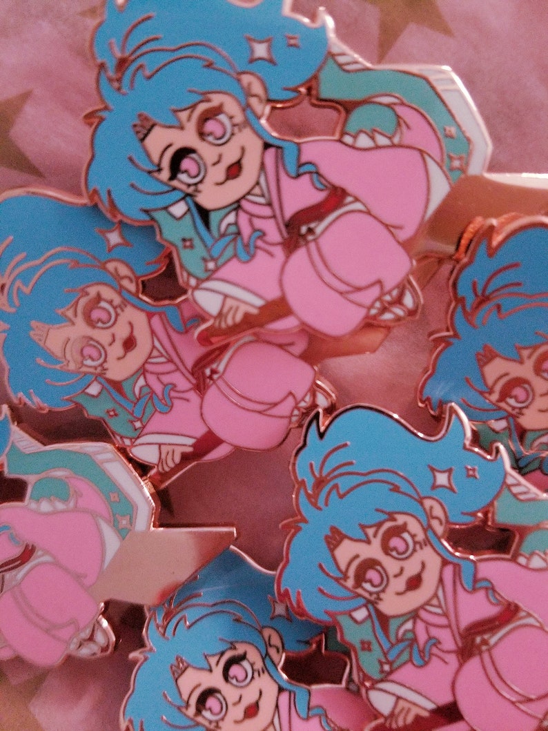 Pu&ograve; includere: Un set di cinque spille smaltate con un personaggio dei cartoni animati con capelli blu, vestiti rosa e occhi grandi. Le spille sono in varie pose e hanno una finitura metallica lucida.