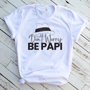 May include: White t-shirt featuring a black and white illustration of a fedora hat and the text "Don't Worry Be Papi" in black and grey lettering.