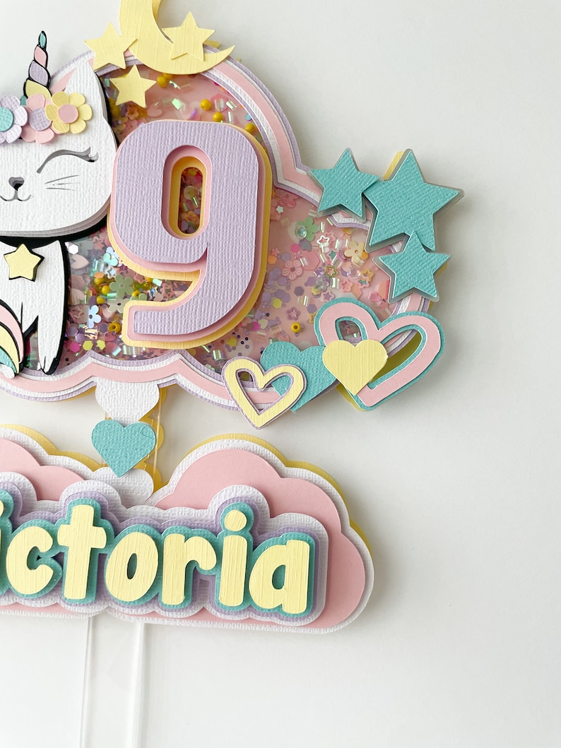 Może przedstawiać: Topper na tort w pastelowych kolorach r&oacute;żowym i ż&oacute;łtym z białym kotem z rogiem jednorożca, cyfrą 9 i imieniem "Victoria" w kształcie chmury.