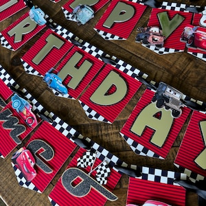 May include: A red and gold "Happy Birthday" banner with a racing theme. The banner features the text "HAPPY BIRTHDAY" in gold letters, with images of race cars and checkered flags. The banner is strung on a black ribbon.