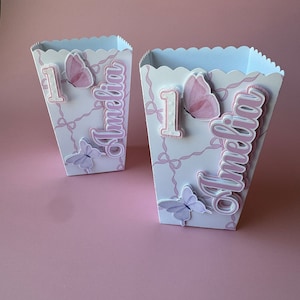 May include: Two white cardboard boxes with a scalloped edge, decorated with pink and white butterflies, bows, and the name "Amelia". The boxes are decorated with the number "1" and are likely for a first birthday party.