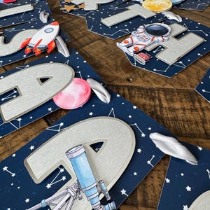 Peut inclure: Décorations de fête sur le thème de l'espace. Elles comprennent des lettres argentées, des fusées, des planètes et un astronaute, sur un fond bleu foncé parsemé d'étoiles blanches, pour une ambiance cosmique.