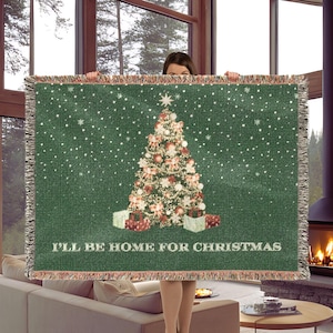 Könnte beinhalten: Eine grüne Webdecke mit einem zentralen, geschmückten Weihnachtsbaum mit Geschenken darunter, umgeben von weißen Schneeflocken, und dem Text "I'LL BE HOME FOR CHRISTMAS" in weißem Druck am unteren Rand, mit Fransenabschluss.