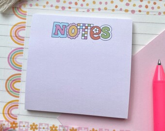 May include: A pink sticky note pad with the word "NOTES" in colorful letters. The pad is surrounded by colorful washi tape and a pink pen.