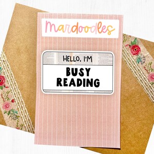 May include: A pink and white striped card with the text "mar doodles" in pink, orange, and yellow lettering. A white sticker with a black border and the text "HELLO, I'M BUSY READING" is on the card.