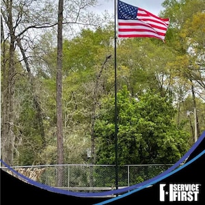 May include: A black telescoping flagpole with an American flag waving in the wind. The flagpole is in front of a green wooded area. The text "SERVICE FIRST" is in the lower right corner of the image.  Telescoping Flagpoles.