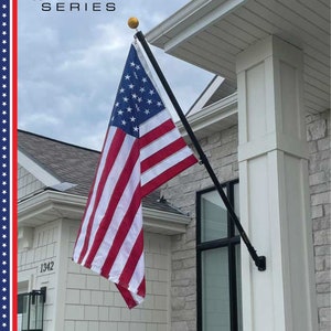 May include: A black flagpole with a gold finial is mounted on the side of a white house. An American flag is flying from the pole. The flag is red, white, and blue with 13 stripes and 50 stars. The house has a white siding and a gray roof. The house number 1342 is visible on the side of the house.