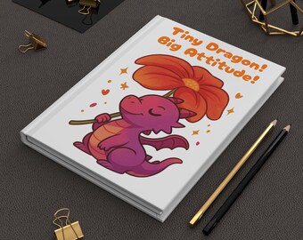 以下が含まれることがあります： 「Tiny Dragon! Big Attitude!」の文字が入った白いジャーナルには、大きなオレンジ色の花を持った漫画のドラゴンが描かれています。ドラゴンは紫とオレンジ色です。金と黒の鉛筆と金のクリップも表示されています。