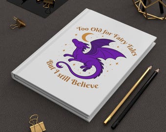 May include: White journal with a purple dragon illustration and the text "Too Old for Fairy Tales But I still Believe". Gold and black pencils and gold binder clips are also visible.