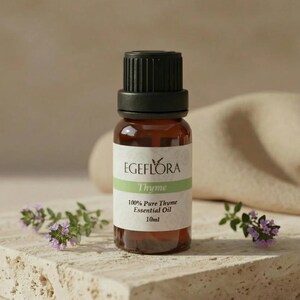 May include: An amber glass bottle of "Egeflora Thyme 100% Pure Thyme Essential Oil 10ml" with a black cap. The bottle is on a stone surface, with sprigs of thyme and purple flowers. The background is a neutral beige.
