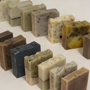 Puede incluir: Una variedad de jabones artesanales en diversos colores y texturas. Los jabones varían desde verde claro y beige hasta marrón oscuro y carbón, con ingredientes naturales visibles. Las barras son rectangulares y están dispuestas en filas, mostrando sus diseños únicos.