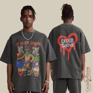 Op de afbeelding: Donkergrijs t-shirt met een afbeelding op de voorkant van een engel met vleugels, een zonnebril en een gezichtsmasker, met de tekst "City of Angels" en "West Coast". De achterkant van het shirt heeft een rood hart met de woorden "Create Thrive".
