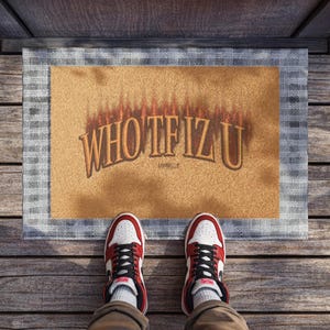 May include: A rectangular doormat with the text "WHO TF IZ U" in a brown, flame-like font. The mat has a tan center and a gray plaid border. Red, white, and black sneakers are visible at the bottom of the image.