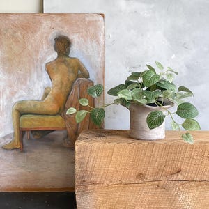 May include: A small potted plant with green and white striped leaves sits atop a weathered wooden block. In the background, a vintage painting depicts a figure seated in a chair, and a gray abstract painting.