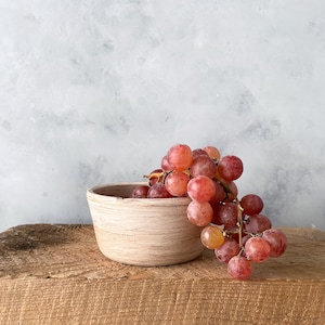 May include: A small, white ceramic bowl with a rustic finish sits on a wooden surface. A bunch of red grapes hangs over the edge of the bowl.